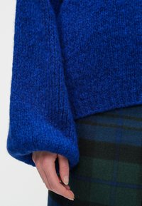 Ręka spoczywa na boku, na sobie ma oversize'owy niebieski sweter i ciemnozieloną spódnicę w niebieską kratę.