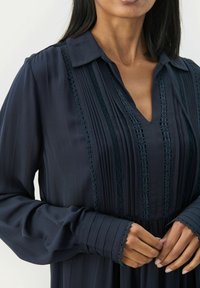 Dunkelblaue Bluse mit V-Ausschnitt, plisseeartigem Design, Spitzenverzierungen und langen Ärmeln mit strukturierten Bündchen. Glatte Stoffqualität mit sanftem Glanz.