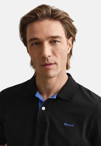 Fekete galléros póló kék gallérral és GANT logóval. Gombos záródással és sima textúrával rendelkezik. Közeli kép a pólóról, semleges háttér előtt.