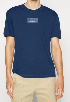 Mężczyzna ubrany w granatowy t-shirt Emporio Armani EA7 i beżowe spodnie, stoi na jednolitym tle.