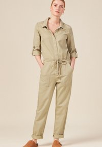 Khaki jumpsuit met een kraaghalslijn, lange mouwen opgerold, een trekkoord in de taille en twee voorzakken. Zachte stof met een gladde textuur.