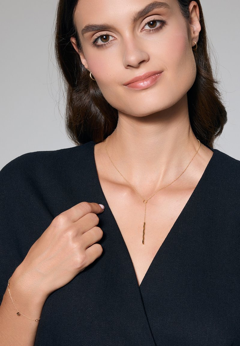 Kobieta z brązowymi włosami do ramion ma na sobie czarną bluzkę, złotą naszyjnik i bransoletkę, delikatnie uśmiechając się w kierunku kamery.