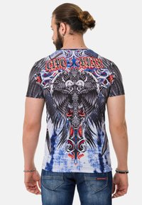 Kurzarm-Grafik-T-Shirt mit lebendigen Schädel- und Adler-Designs in Schwarz, Rot und Blau auf einem weißen Hintergrund mit einem Batik-Effekt.