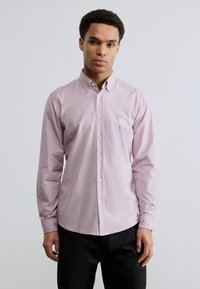 Νεαρός άνδρας στέκεται, ντυμένος με ανοιχτό ροζ πουκάμισο με κουμπιά και μικρό λογότυπο "BOSS" στο στήθος και μαύρο παντελόνι, μπροστά σε απλό λευκό φόντο.