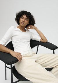 Woman with curly hair wearing a white top and beige pants seated on a modern black chair with armrests against a plain background.