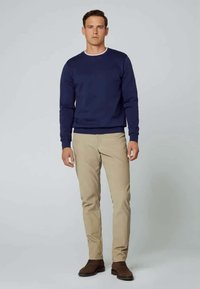 Sweatshirt azul-marinho com um decote canelado, combinado com calças chino bege. Modelo a usar sapatos castanhos está posicionado contra um fundo cinza-claro.