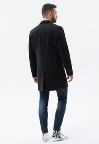 Schwarzer Mantel mit langer Silhouette, mit Reverskragen und Knopfakzenten. Getragen über blauen Jeans und weißen Sneakern mit grafischem Detail.