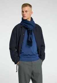 Marinblå jacka med elastiska manschetter, blå tröja och en mörkblå scarf med fransar. Grå byxor fulländar outfiten.