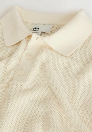 Krémová polo košeľa s textúrovaným pletením, s klasickým golierom a trojicou gombíkov. Značka je viditeľná vo vnútri goliera.