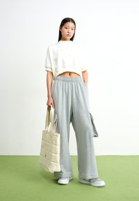 Camisola branca curta com logótipo "NIKE", combinada com calças de fato de treino cinzentas largas com bolsos laterais. Saco tote bege acolchoado. Piso verde.