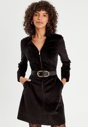Robe en velours côtelé noir avec une fermeture éclair sur le devant, des manches longues et des poches latérales, cintrée à la taille avec une ceinture noire dotée d'une boucle en or ronde.