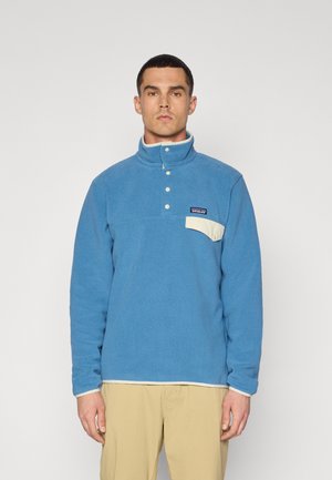 Niebieski polarowy sweter z wysokim kołnierzem, zapięciem na zatrzaski z przodu oraz kontrastową kremową kieszenią. Posiada małe logo na piersi.
