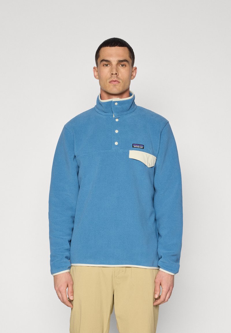 Blå fleece-pullover med høj krave, fremadgående trykknaplukning og en kontrasterende cremefarvet lomme. Har et lille logo på brystet.