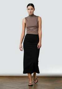 Brunærmeløs mock neck top i tætsiddende design. Sort, ankel-længde nederdel med lige silhuet. Model iført flip-flops på et trægulv.