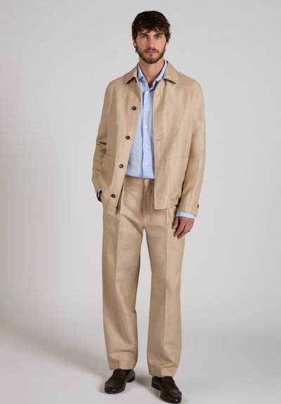 Hombre vistiendo chaqueta y pantalones beige, camisa de botones azul claro y zapatos marrón oscuro, de pie contra un fondo gris liso.