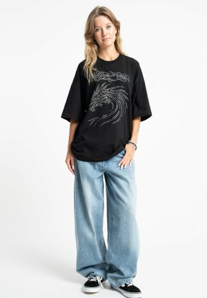 Młoda kobieta ubrana w czarny, oversize'owy t-shirt z motywem smoka i luźne jasnoniebieskie jeansy stoi na jednolitym białym tle.