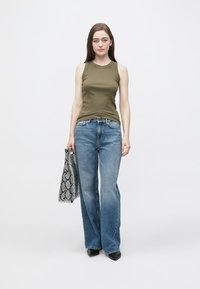 Top verde oliva senza maniche, jeans blu a zampa d'elefante e scarpe nere con punta; tiene in mano una borsa nera e bianca con motivo a serpente.