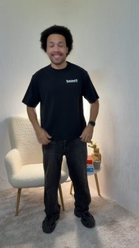 Mladý muž s kučeravými vlasmi v čiernom tričku Timberland, tmavých džínsoch a čiernych topánkach stojí v miestnosti s bielou štruktúrovanou stenou a kreslom.