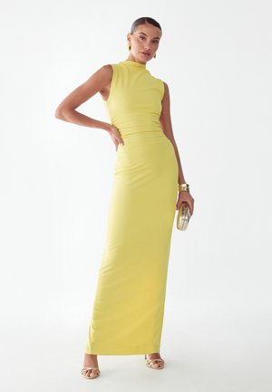 Vrouw draagt een mouwloze, citroengele, nauwsluitende maxi-jurk met gouden oorringen, armbanden, sandalen met bandjes en houdt een gouden clutch vast.