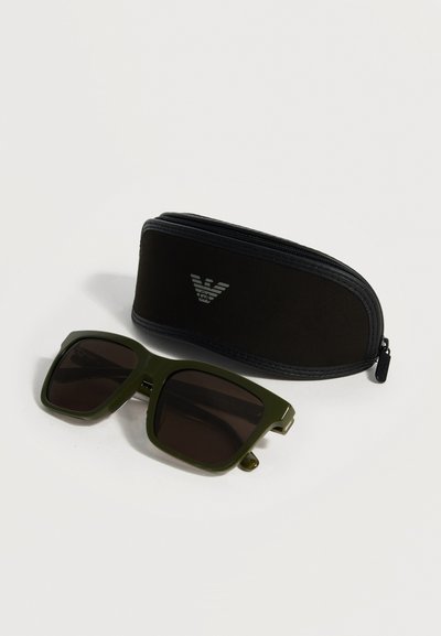 Donkergroene vierkante zonnebril met zwarte glazen naast een zwarte etui met ritssluiting met een zilveren logo op een witte achtergrond.