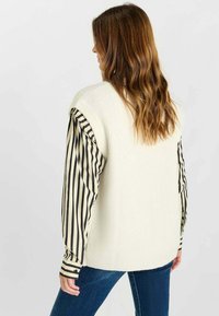 Maglione smanicato color crema a costine indossato su una camicia a maniche lunghe bianca e nera a righe, abbinato a jeans di denim blu, che mostrano una vestibilità comoda.
