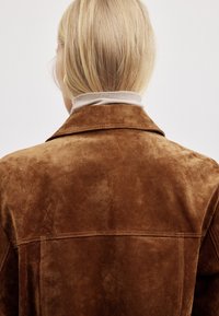 Brun ruskindsjakke med en glat tekstur, der har en standardkrave og synlige syninger. Båret over en beige turtleneck.