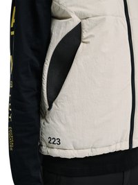 Beżowa pikowana kamizelka z czarną siateczkową kieszenią na zamek, numer "223" nadrukowany blisko dołu, noszona na czarnej koszulce z długim rękawem z żółtym tekstem na rękawie.