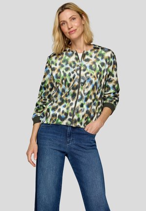 Frau mit schulterlangem blondem Haar, die eine mehrfarbige Leopardenprint-Jacke mit Reißverschluss und blaue Jeans trägt, mit einer Hand in der Tasche stehend.