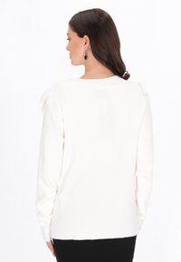 Biały sweterek dzianinowy z okrągłym dekoltem, długimi rękawami i falbanami na ramionach. Miękka faktura, luźny krój i długość do bioder.