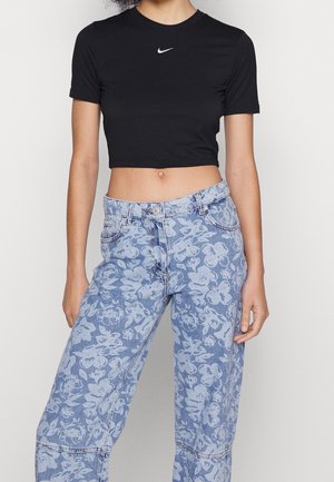 Femme portant un t-shirt court Nike noir et un jean bleu taille haute avec un motif floral blanc, debout devant un fond blanc.