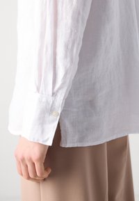 Witte linnen shirt met een relaxte pasvorm, voorzien van een knoopmanchet, subtiele kreuktextuur en zijsplitten, gecombineerd met beige broek.