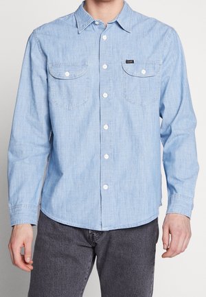 Mann trägt hellblaues Hemd mit zwei Knopfleisten auf der Brust und dunkelgraue Hose, steht vor einfachem Hintergrund.