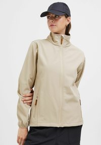 Beige softshell jacket with a high collar, zippered pockets, and a smooth texture. Brown accents at the zippers. Designed for outdoor use.