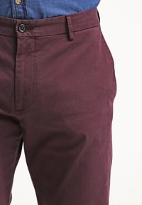 Pantaloni di cotone bordeaux con chiusura a bottone, caratterizzati da una texture liscia e tasche laterali. Parzialmente visibili in vita.