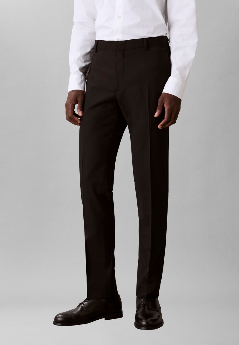 Man wearing black tailored trousers, black leather shoes, and a white dress shirt against a plain light gray background.