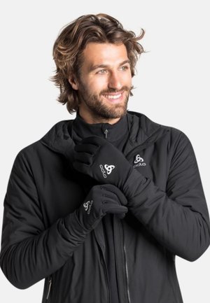 Mann trägt schwarze Zip-up-Jacke und passende Handschuhe mit sichtbarem Odlo-Logo, lächelt und schaut zur Seite.
