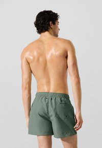 Zelené plavecké šortky s elastickým pásom, zadným vreckom a hladkou, mierne lesklou textúrou. Dizajn je jednoduchý a funkčný.