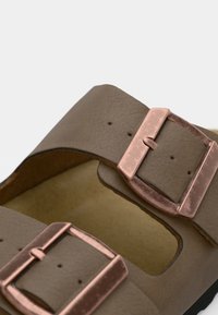 Brun sandalrem med två stora roséguldfärgade metallspännen och flera justeringshål på en ljus bakgrund.