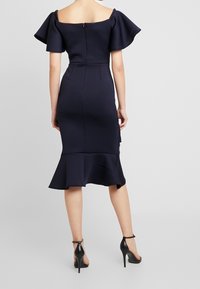 Marinblå klänning med off-the-shoulder design och svängda ärmar, kroppsnära passform, dragkedja bak och volangkant. Bärs med svarta högklackade skor.