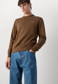 Bruine gebreide trui met een ronde hals, geribde manchetten en tailleband. Gecombineerd met blauwe denim jeans met een rechte pijp.