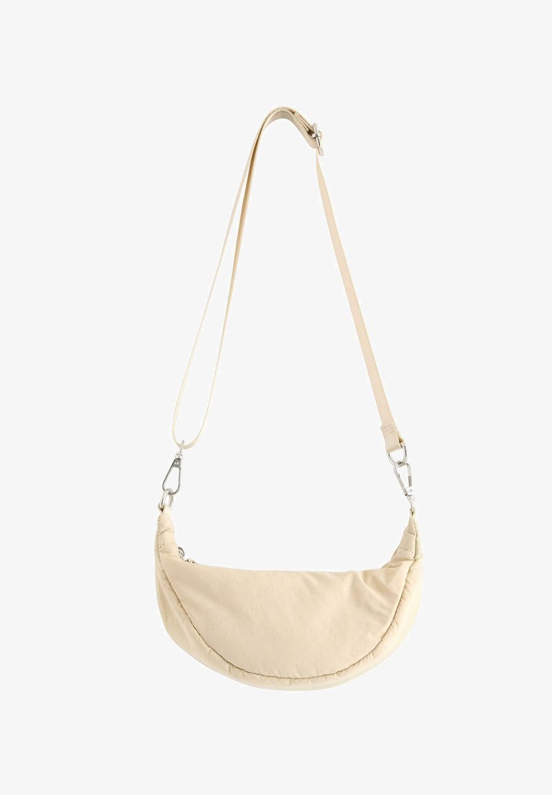 Beige skuldertaske i halvmåneform med justerbar rem og sølvsfarvede metalspænder på en hvid baggrund.