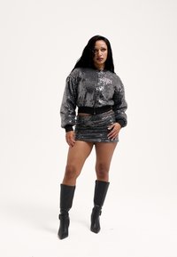 Fekete flitteres bomber dzseki cipzárral és logóval. Egy hozzáillő flitteres miniszoknyával és térdig érő fekete csizmával párosítva.