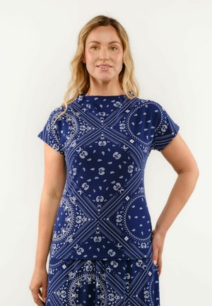 Frau trägt ein marineblaues kurzärmeliges Oberteil mit weißen Paisley- und geometrischen Mustern, steht mit einer Hand in der Hüfte.