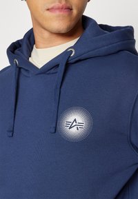 Homme portant un sweat à capuche bleu marine avec un logo circulaire blanc arborant un "A" stylisé et des rayures sur la poitrine, sous une chemise crème.