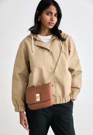 Kvinde iført beige hættetrøje og sorte bukser med en brun skuldertaske med guldkæde og lås.