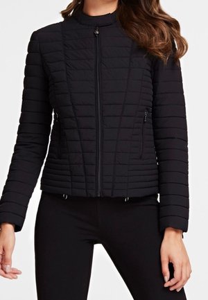 Vrouw draagt een nauwsluitend, doorgestikt zwart jack met een ritssluiting aan de voorkant en ritssluitingen op de zijzakken, gecombineerd met zwarte broek.