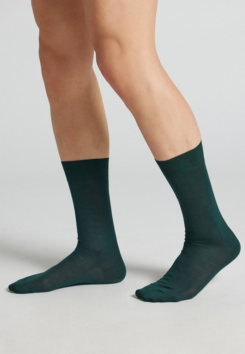 Gambe con calzini verde scuro a metà polpaccio, in piedi su una superficie grigio chiaro con uno sfondo semplice.