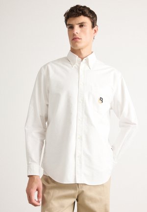 En ung mann iført en hvit skjorte med knapper og et lite brodert logo på brystlommen, samt beige bukser, står med den ene hånden i lommen.