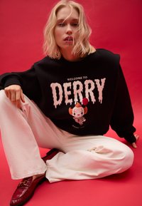 Felpa nera con stampa "WELCOME TO DERRY" e personaggio dei cartoni animati. Abbinata a pantaloni larghi bianchi e mocassini rossi. Sfondo rosa.