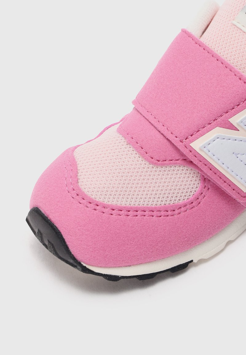Zapatilla deportiva rosa con una parte superior de ante y malla, con una correa de velcro y acentos blancos. Suela de goma negra con relieve texturizado.